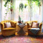 Un acogedor rincón de sala de estar con dos sillones de terciopelo amarillo, una pequeña mesa de madera y una variedad de plantas verdes colgantes y en macetas frente a una ventana cubierta por cortinas con dibujos rojos.