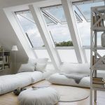 Un dormitorio abuhardillado minimalista y bien iluminado con tragaluces, una cama baja con ropa de cama blanca, una alfombra blanca y una estantería junto a una escalera de metal con alternativas al aire acondicionado para mantener