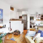 Se está reformando una sala de estar acogedora y bien iluminada con una cocina abierta, con electrodomésticos modernos, una guitarra en la esquina, almohadas decorativas en el sofá y varias obras de arte en las paredes.