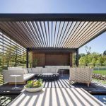 Un moderno espacio al aire libre con un juego de sofás blancos, una mesa de café y sillones, con detalles de listones de madera que brindan sombra parcial desde una pérgola bioclimática, ubicada en