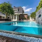Una moderna piscina de verano con una pared transparente, situada en el patio trasero junto a una casa elegante y rodeada de una cubierta de madera y pais.