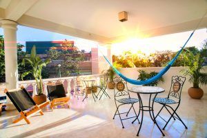 Una terraza soleada de verano con una hamaca azul, sillas reclinables y una mesa con sillas decoradas y rodeadas de plantas en macetas durante el año.
