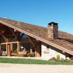 Una casa rústica moderna, frente a un cielo azul claro, refleja el sueño de una casa reformada con su gran techo voladizo, vigas de madera a la vista y una chimenea de piedra.