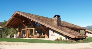 Una casa rústica moderna, frente a un cielo azul claro, refleja el sueño de una casa reformada con su gran techo voladizo, vigas de madera a la vista y una chimenea de piedra.