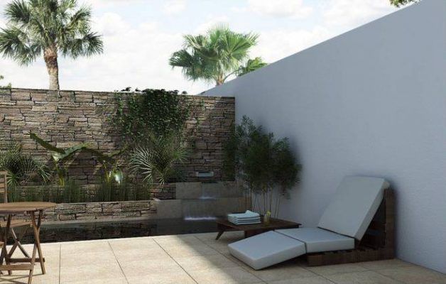 Un espacio exterior moderno con un muro de piedra con vegetación, una mesa de madera, un jardín sin mantenimiento con una tumbona y un libro encima, y palmeras contra un cielo despejado.