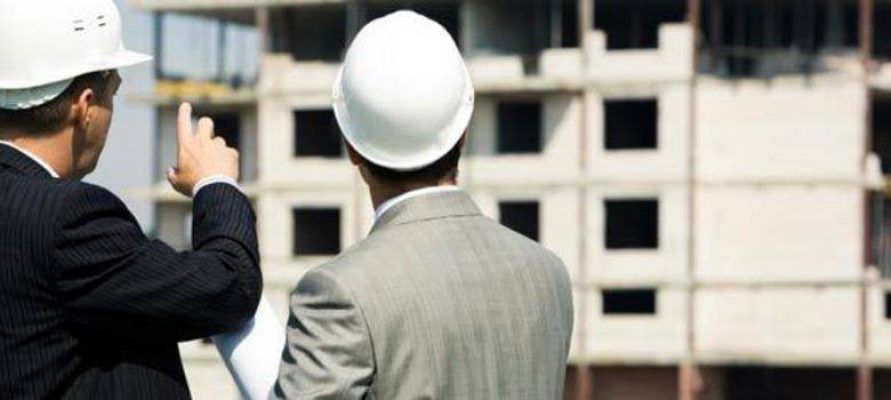 Dos arquitectos con cascos están examinando un sitio de construcción; uno está haciendo gestos hacia el edificio.