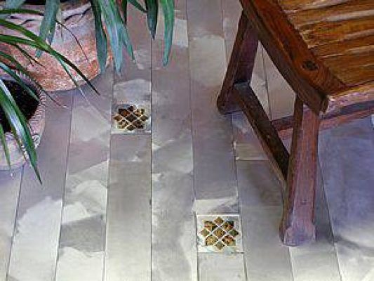 Suelo de baldosas con inserciones metálicas cerca de la pata de una silla de madera y el follaje de una planta.
