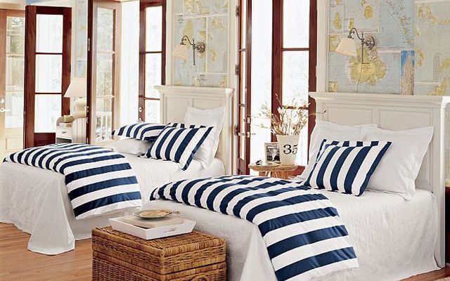 Una casa con un dormitorio bien iluminado que cuenta con dos camas individuales con ropa de cama blanca y detalles de rayas azules, una mesa de mimbre,