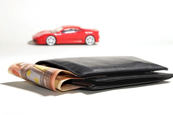 Sobre una superficie hay una cartera negra llena de billetes en euros y al fondo un coche de juguete rojo, que simboliza el sueño de alquilar una furgoneta.
