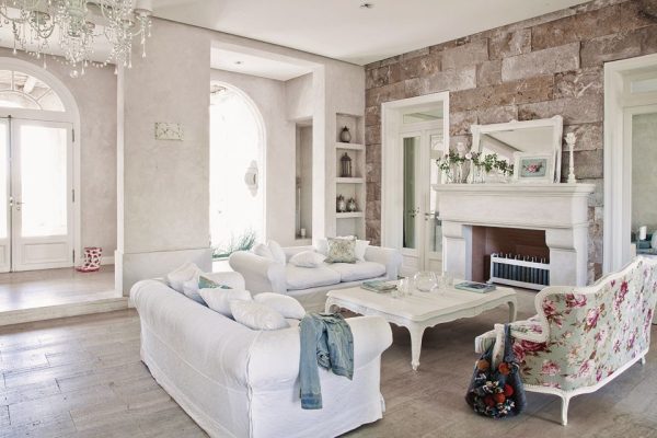 Elegante sala de estar con sofás blancos, un sillón floral y una chimenea clásica, acentuada por luz natural y candelabros, que muestra el Estilo Shabby chic.