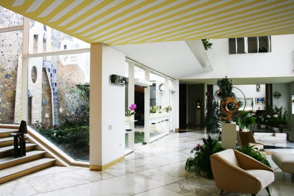 Vestíbulo moderno con techo a rayas, una variedad de plantas y una decoración contemporánea inspirada en Vivir a lo Gio Ponti, con una pequeña fuente de agua interior visible al fondo.