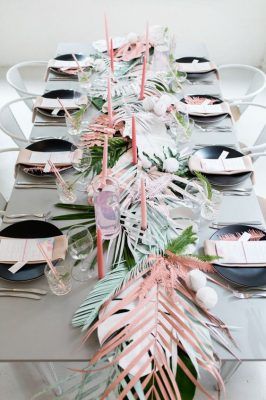 Una mesa está preparada para una comida formal con vajilla moderna y decorada con un ambiente navideño que incluye velas rosas, flores blancas y rosadas y hojas de palma pintadas.