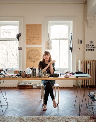 Una mujer sentada en una silla en un estudio bien iluminado, rodeada de materiales de arte y notas sobre "reformas integrales" adheridas a la pared detrás de ella.