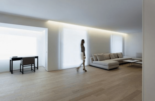 Una persona camina en un salón moderno iluminado brillantemente, con mobiliario mínimo que incluye un sofá, un escritorio con una silla y una factura de