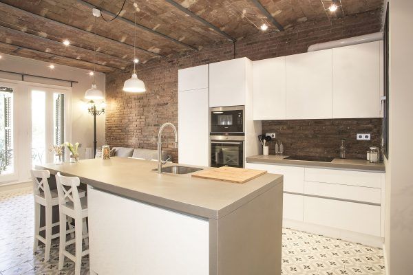 Una cocina moderna con gabinetes blancos, electrodomésticos de acero inoxidable y una isla central con asientos, apoyada contra una pared de ladrillo visto, todo parte de una reforma integral.