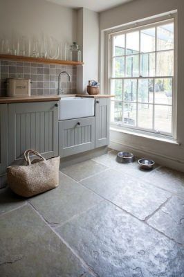 Un diseño de cocina ordenado con suelo de baldosas de piedra, gabinetes grises, una ventana con vistas al exterior, estantes abiertos con cristalería y