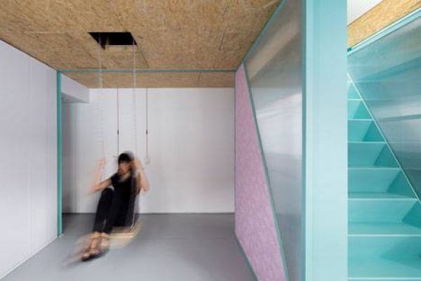 Una persona está sentada en un columpio en una habitación moderna y minimalista con paredes grises, una escalera de color agua y un techo de madera durante un recorrido por la casa.