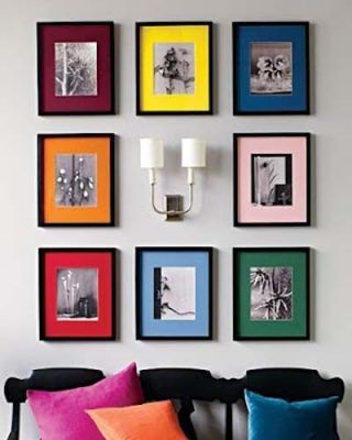 Una colección de fotografías en blanco y negro expuestas en coloridos marcos sobre una pared gris, flanqueada por una lámpara de pared de doble brazo y real.