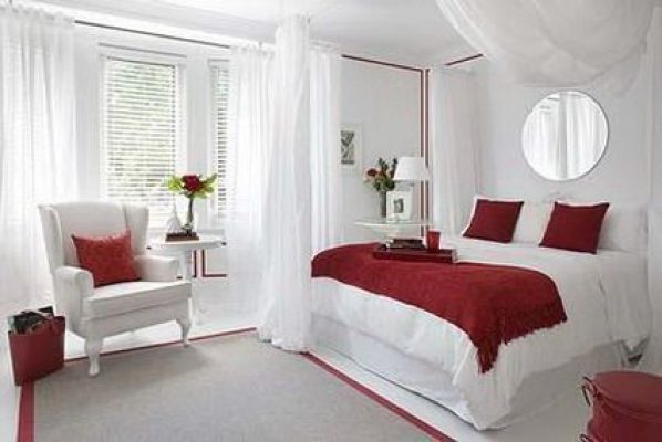 Un dormitorio cuidadosamente arreglado con paredes blancas, diseñado según los principios del Feng Shui, con una cama blanca con detalles en rojo, un sillón blanco a juego, una mesa auxiliar y un espejo redondo, acentuado por