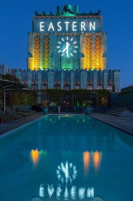 Un edificio iluminado con la palabra "este" en su fachada se eleva sobre una tranquila piscina en la azotea durante el crepúsculo en Los Ángeles.