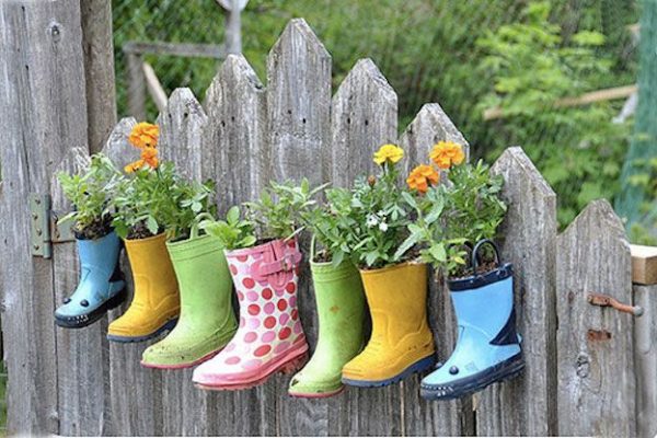 Viejas botas de goma de colores reutilizadas como jardineras para un huerto urbano, colgadas de una valla de madera.