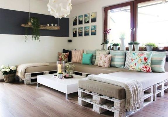 Una acogedora sala de estar que presenta muebles hechos de palés de madera reciclada, con cojines en diversos patrones y colores, iluminación ambiental.