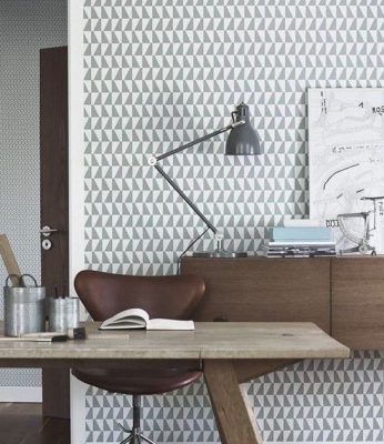 Un rincón de oficina en casa con un diseño de papel tapiz geométrico, que presenta un escritorio de madera reformado, una lámpara de escritorio ajustable negra y una silla de cuero. Hay libros y artículos de papelería sobre el escritorio.