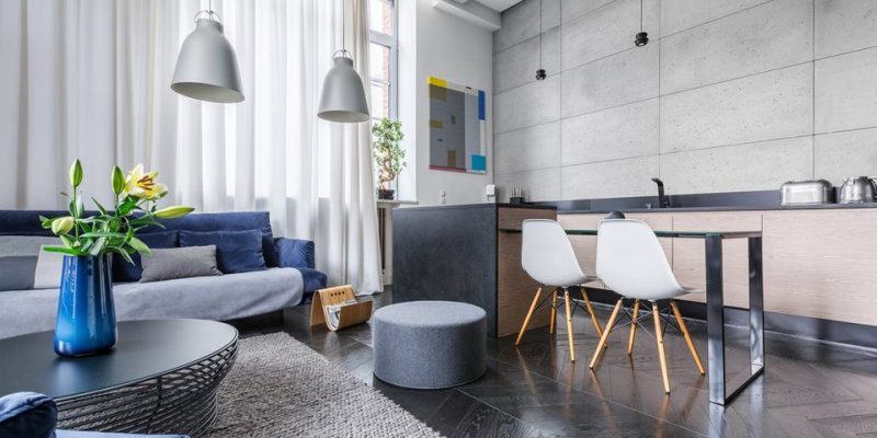 Una sala de estar moderna con un sofá gris, un cojín decorativo en azul, un jarrón azul con flores sobre una mesa de café redonda oscura y un área de comedor con sillas blancas y una encimera de cocina al fondo.