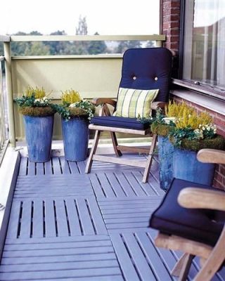Una acogedora terraza con piso de madera cuenta con un sillón reclinable de color azul oscuro y dos grandes maceteros azules con plantas verdes, todo ello frente a una barandilla y una vista del edificio adyacente.