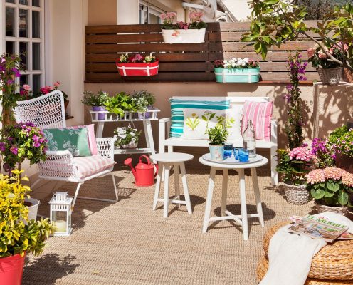 Un colorido y acogedor pequeño paraíso al aire libre con una mesa blanca, dos sillas con cojines rosados, plantas en macetas, flores y
