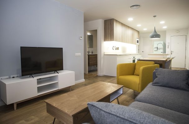 Una sala de estar moderna se transforma perfectamente en un área de cocina, con paredes neutras, un sofá gris, un sillón amarillo, decoración de salones con un televisor de pantalla plana sobre una unidad de entretenimiento blanca.