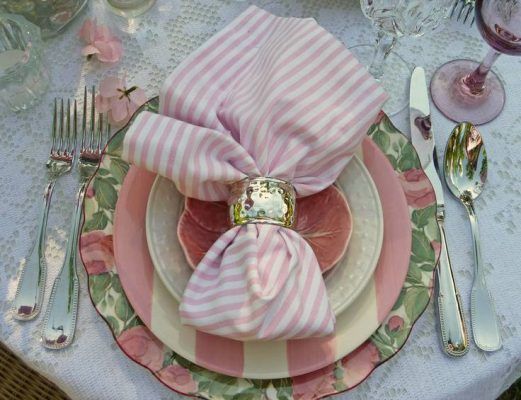 Una mesa elegantemente decorada con un tema floral, presentando una servilleta rosa y blanca a rayas doblada y sostenida por un anillo encima de platos.