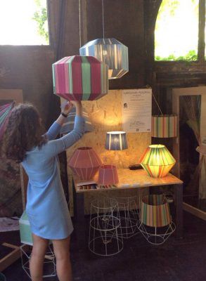 Una persona está ajustando una colorida lámpara colgante con forma geométrica en una tienda emergente con varias lámparas en exhibición.