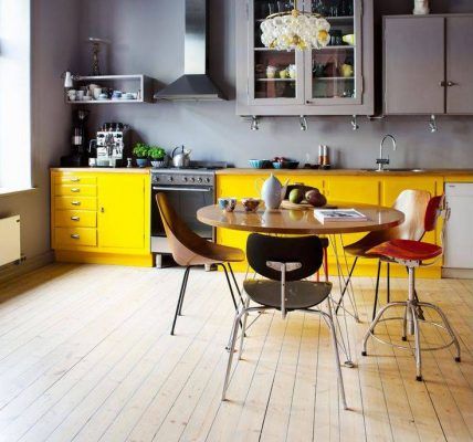 Una cocina contemporánea con gabinetes amarillos, una mesa de comedor central con sillas mezcladas y estantes superiores grises contra una pared neutra, presentando