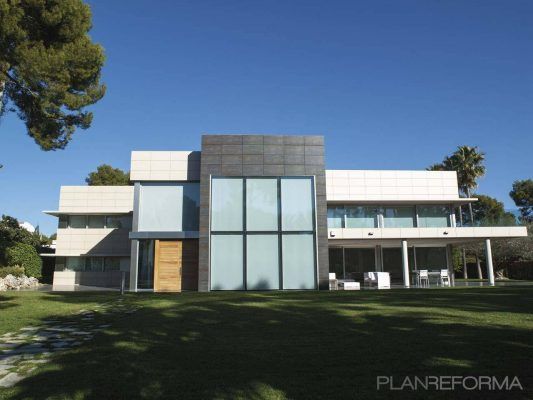 Una casa moderna con grandes ventanales, techos planos y una combinación de materiales de hormigón, vidrio y madera, rodeada por un césped bien cuidado con árboles altos al fondo. el crecen