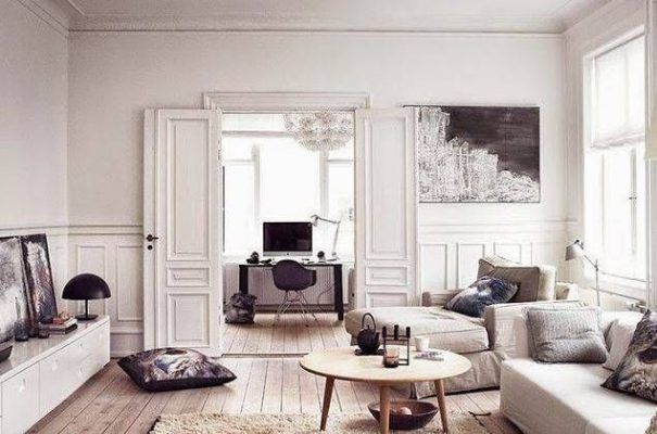 Una sala de estar en tonos neutros con un sofá blanco de moda, una mesa de café redonda con elementos decorativos, cojines en el piso, una silla y un escritorio negros con una computadora y obras de arte sobre una clásica cama doble blanca.