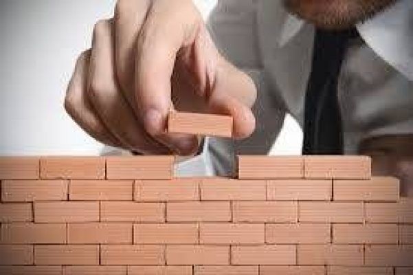 Una persona con camisa coloca un bloque de madera encima de una pared de ladrillos cuidadosamente apilados, simbolizando la construcción o la planificación cuidadosa.