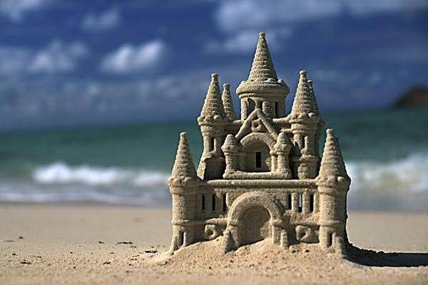 Intrincado castillo de arena con múltiples torretas y arquitectura detallada en una playa de arena, con el mar y un cielo nublado al fondo.