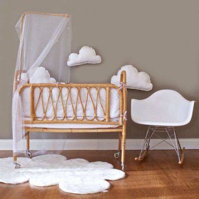 Una cuna de madera antigua con un dosel transparente de inspiración decorativa se coloca junto a una mecedora moderna blanca en una habitación infantil con decoraciones en forma de nubes y detalles de alfombras blancas y esponjosas en el piso.