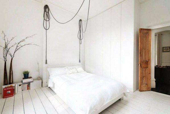Una habitación minimalista con paredes blancas, una colcha blanca sobre la cama, suelo de madera y una única rama como decoración en un jarrón.