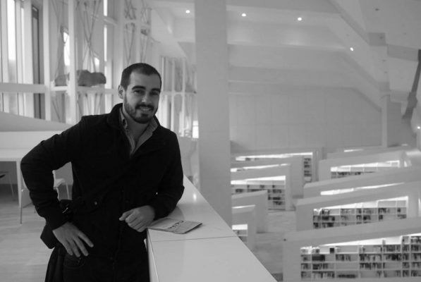 Un hombre con barba y abrigo oscuro está apoyado en una barandilla de la moderna biblioteca del Colegio de Interioristas de Asturias, con estanterías al fondo. la foto es