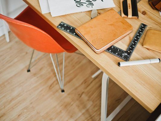 Un espacio de trabajo ordenado con un escritorio de madera con una libreta marrón cerrada, una regla y un marcador negro, junto a una silla roja con patas de metal, encarna claves de éxito para cualquiera que busque empre