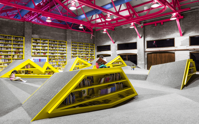 Una biblioteca y centro cultural de interiorismo comercial en Monterrey, con vibrantes estantes amarillos con forma de pirámides, vigas rosadas en el techo e individuales.