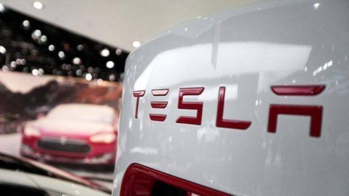 Primer plano de la parte trasera de un vehículo Tesla blanco con el logotipo de Tesla, con otro coche Tesla rojo con baterías de Tesla y un cartel promocional difuminado al fondo.
