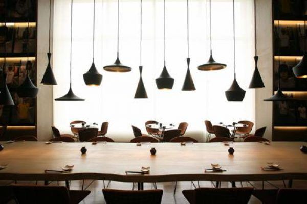 El interior de un restaurante moderno con una larga mesa común, sillas a juego y una serie de lámparas colgantes Tim Dixon Beat, colocadas contra una pared brillante y retroiluminada.