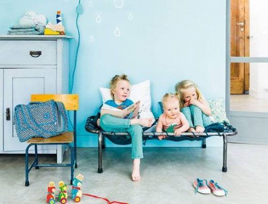 Tres niños sentados en un banco contra una pared azul con patrones de gotas de lluvia, con juguetes esparcidos en el suelo de una habitación