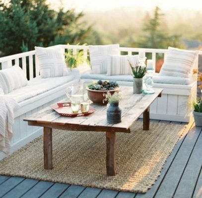 Una zona para sentarse al aire libre sobre una terraza de madera con una mesa rústica, bancos acolchados y una alfombra texturizada, con un telón de fondo de árboles bajo el suave resplandor del atardecer, prepara para la terraza verano.