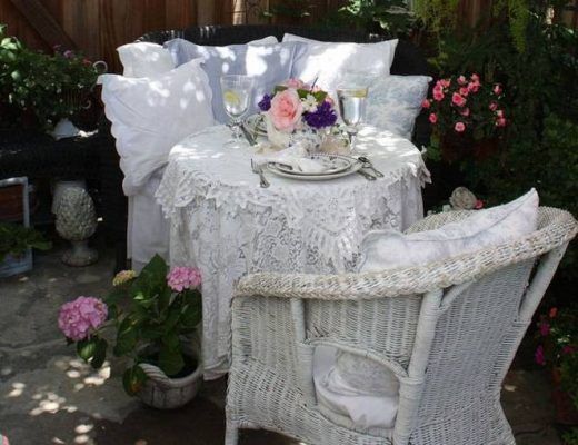 Un pintoresco comedor al aire libre Shabby Chic con una mesa cubierta de encaje para dos personas, vajilla elegante y un jarrón de flores frescas, rodeado de exuberantes plantas de jardín.