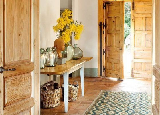 Un recibidor rústico con puertas de madera y suelo, decorado con una mesa auxiliar que tiene un jarrón de flores amarillas, frascos.