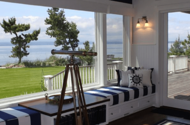 Un acogedor rincón junto al mar con un asiento incorporado junto a la ventana adornado con cojines de temática náutica, inspiración marinera y un telescopio antiguo con vista al océano.